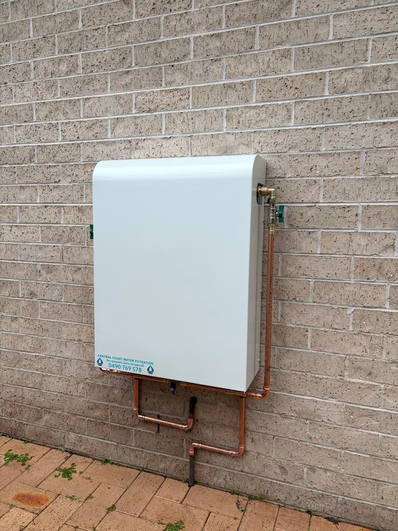 White whole house water filtration system mounted on a face brick wall above paving, with tidy copper pipework and a bypass underneath