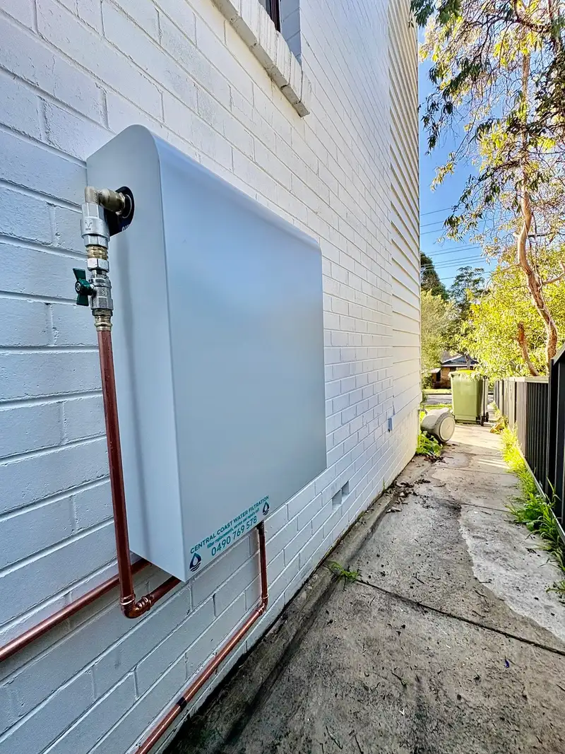 White whole house water filtration system mounted on a painted brick wall along a side path, with copper pipework and an isolation valve