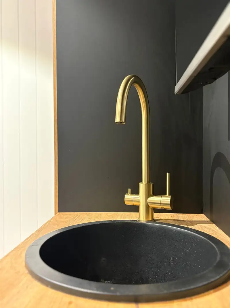 Brushed brass three-way kitchen tap with separate handles for filtered and mixed water, over a round black sink