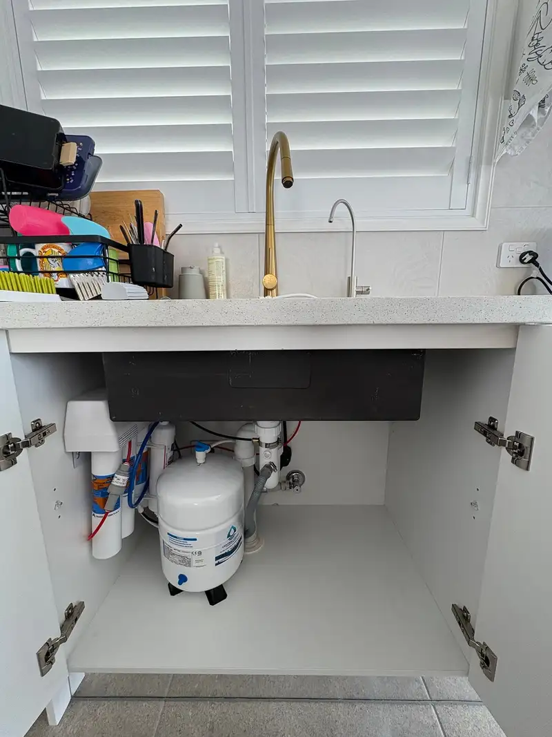 Open kitchen sink cupboard with a reverse osmosis system and storage tank tucked to the left, below a brass mixer and brushed filtered water tap