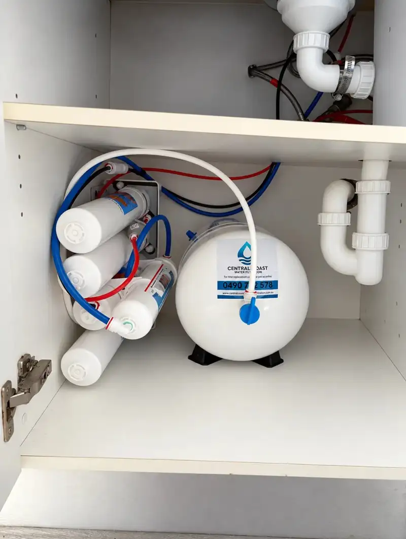Reverse osmosis system mounted sideways with its storage tank in the bottom of a kitchen cupboard, below a shelf and the sink plumbing