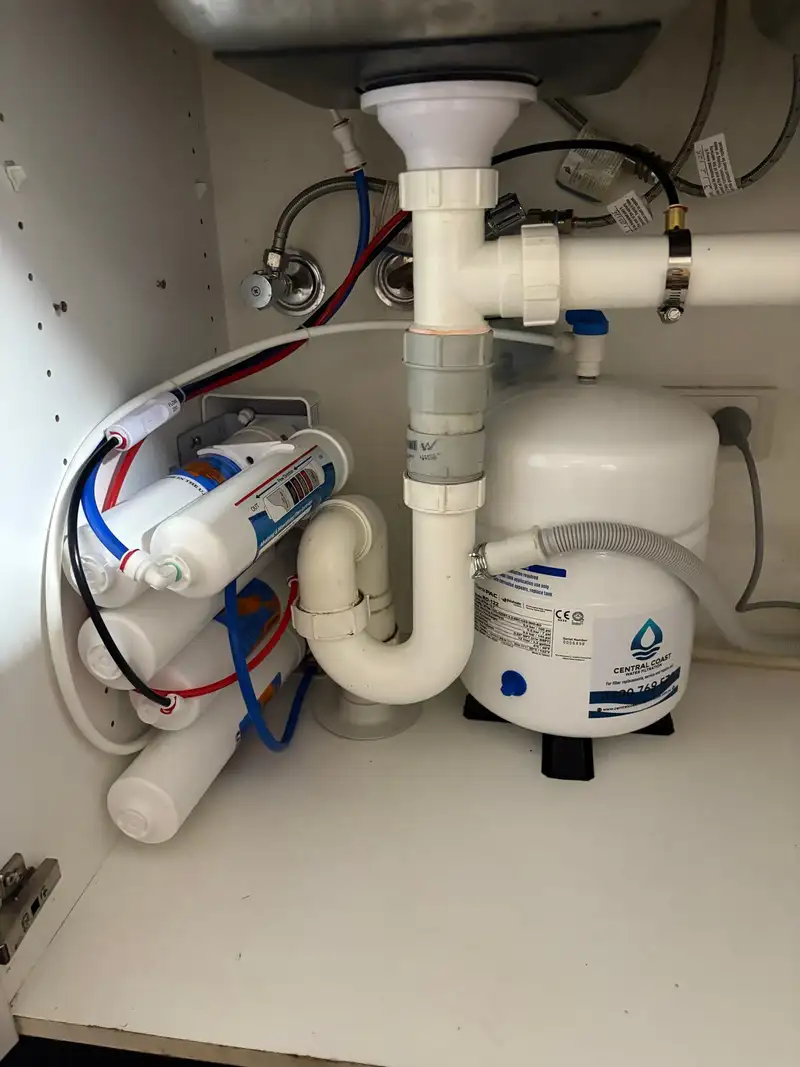 Reverse osmosis system mounted sideways under a kitchen sink next to its storage tank, around the sink's waste plumbing