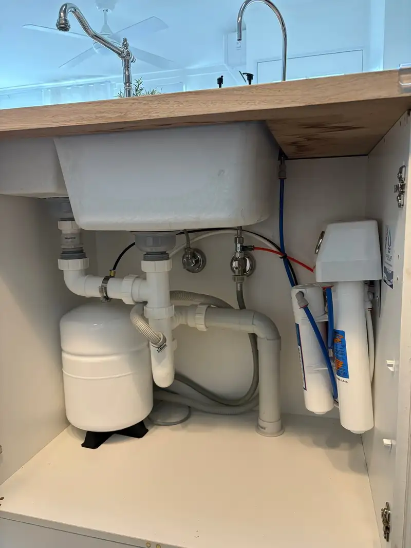 Under-sink cupboard with a reverse osmosis unit on the right-hand wall and its storage tank in the left corner, below polished taps